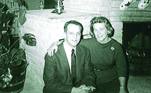 Marjorie and Ernest Flegenheimer in 1962 at home in Green Bay, Wisconsin. Brick fireplace in background and plant to the left. Marjorie and Ernest are seated on the fireplace hearth dressed in formal attire. Marjorie has her right arm around Ernest’s shoulder.